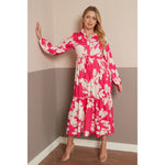 A woman models a long-sleeved midi dress with a vibrant pink and cream floral pattern, standing in a two-toned beige and gray room corner.