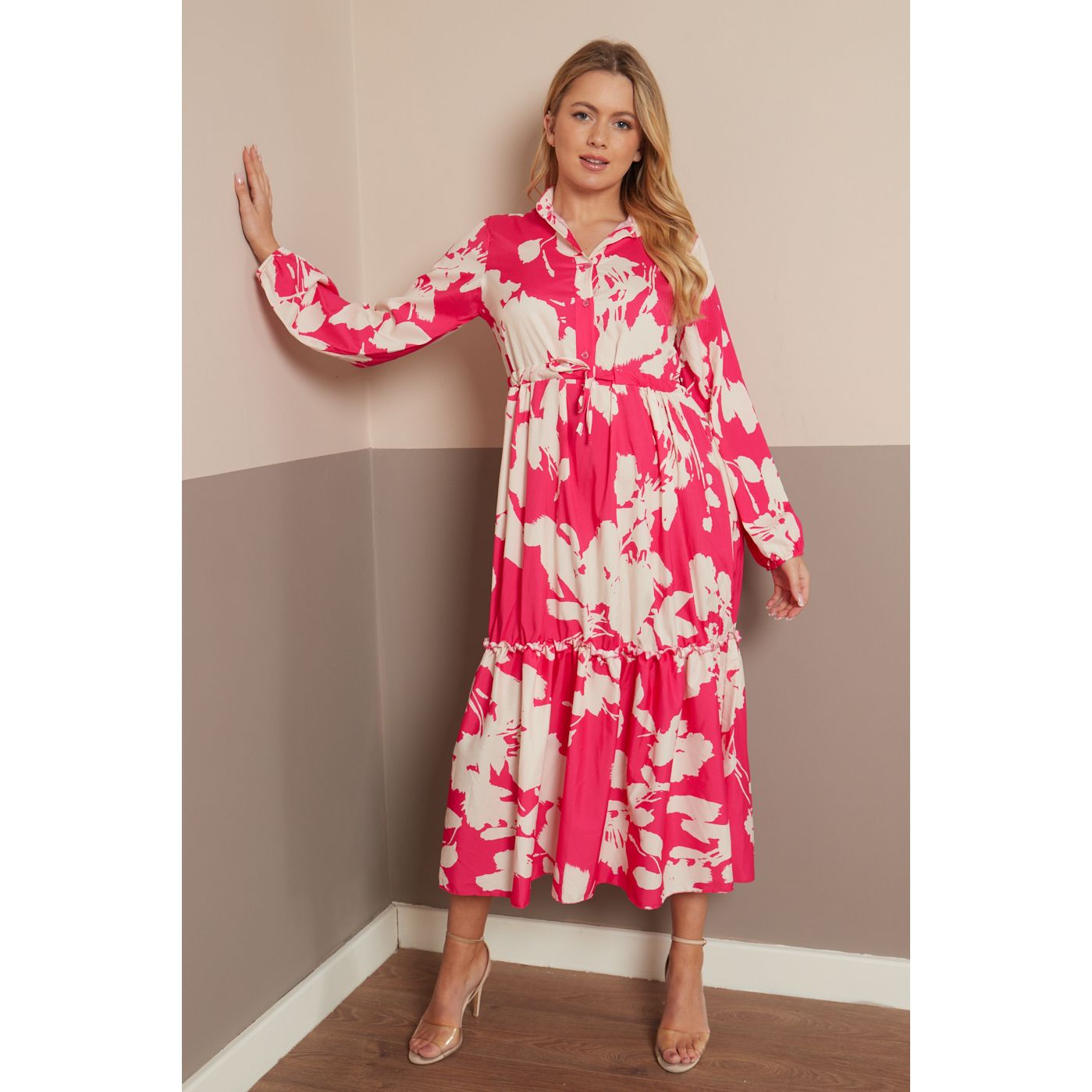 A woman models a long-sleeved midi dress with a vibrant pink and cream floral pattern, standing in a two-toned beige and gray room corner.