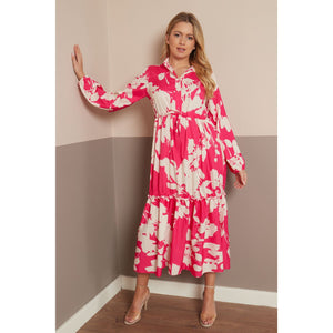 A woman models a long-sleeved midi dress with a vibrant pink and cream floral pattern, standing in a two-toned beige and gray room corner.