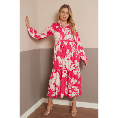 A woman models a long-sleeved midi dress with a vibrant pink and cream floral pattern, standing in a two-toned beige and gray room corner.