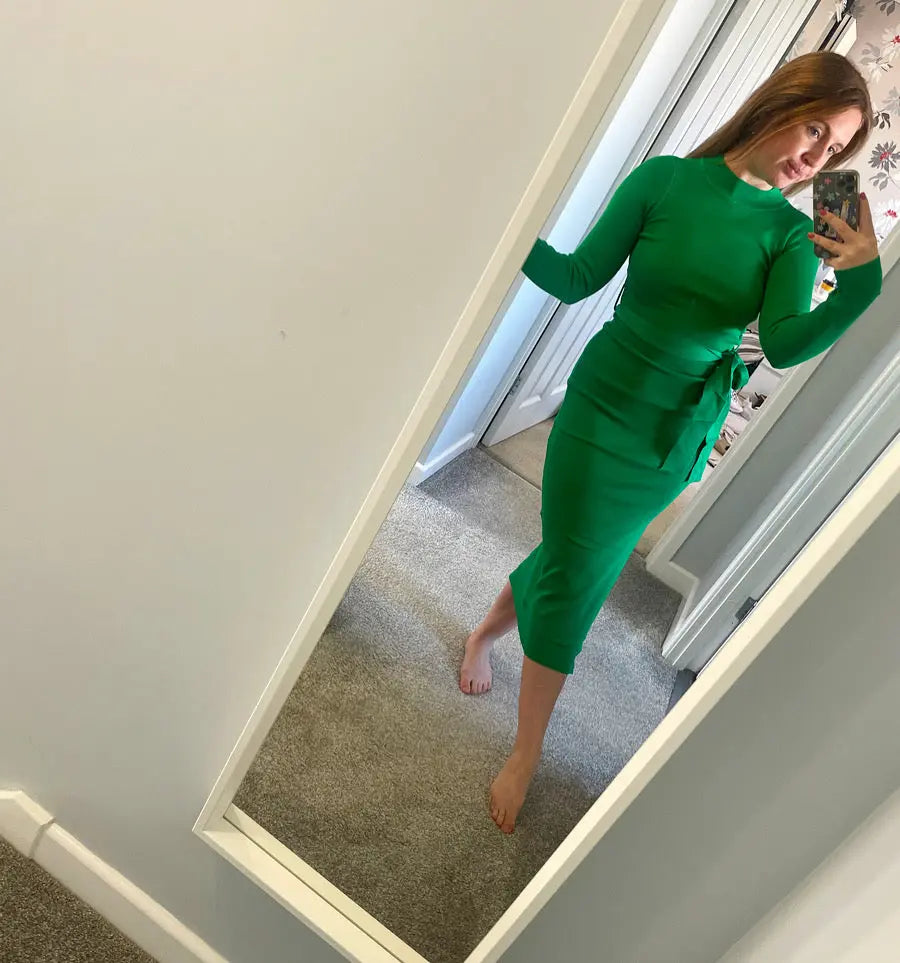 A woman takes a mirror selfie wearing a bright green long-sleeve midi dress with a tie-waist detail, standing in a home hallway.
